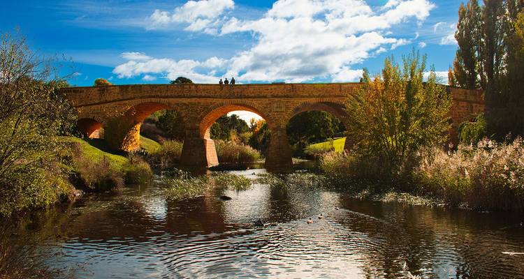 Pont en arche de grès historique enjambant une rivière paisible avec le feuillage automnal et un ciel bleu éclatant.