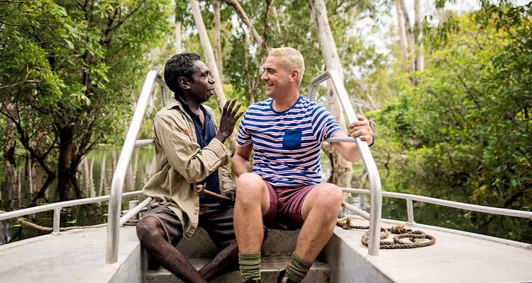 Two men sit on a small boat surrounded by lush wetland forest, chatting and smiling.