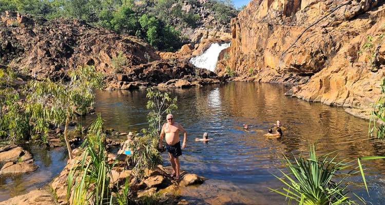 Travelers swim and rest by a natural rock pool beneath a small waterfall in rugged bushland.