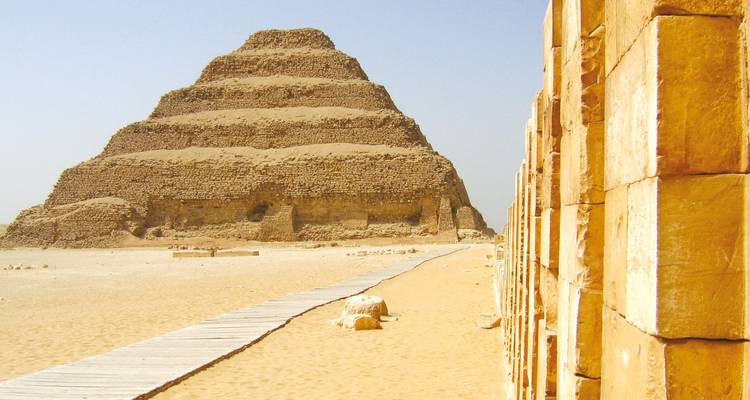 Ancient Step Pyramid of Saqqara rises from the sandy desert with a wooden walkway leading toward it.