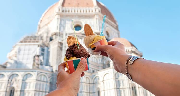 Twee handen die kleurrijke gelato-bekers met wafelbeschuitjes omhoog houden voor Florence's iconische Duomo onder een heldere blauwe hemel.