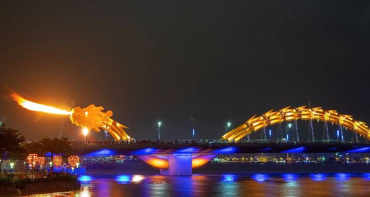 Menschenmengen beobachten, wie die Drachenbrücke von Da Nang Feuer über den Fluss speit, umgeben von farbenfroher blauer und oranger Nachtbeleuchtung.