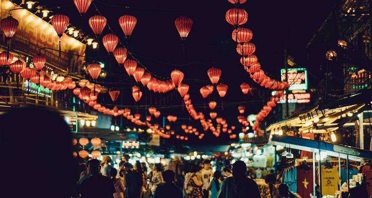 Mercado nocturno vibrante iluminado por filas de faroles rojos con multitudes explorando puestos.