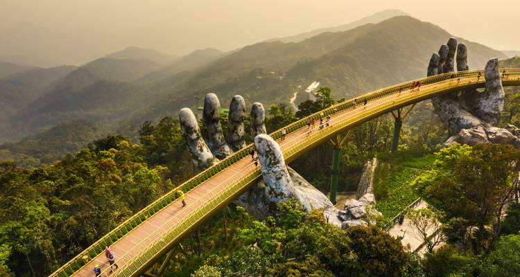 Pont doré tenu par des mains de pierre géantes dans un paysage montagneux.