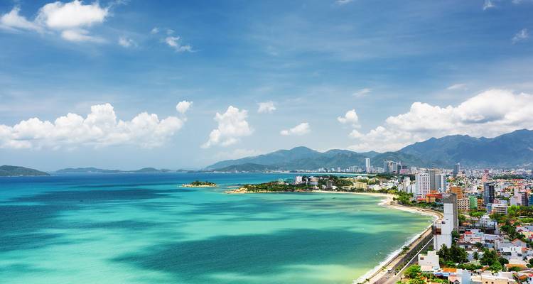 Vue aérienne panoramique d'une baie turquoise, d'un littoral aux sables blancs et d'une ville côtière moderne encadrée par des montagnes lointaines.