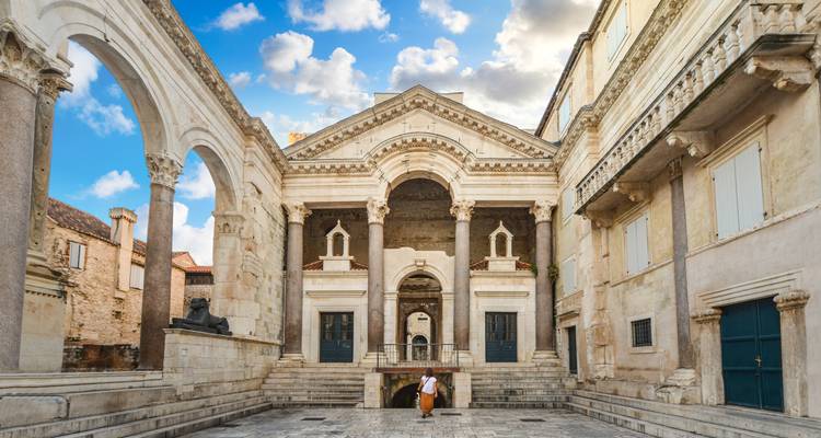 Un musicien se tient sous les arcades ornées de la cour du Péristyle de Split, encadré par un ciel bleu et des colonnes en marbre.