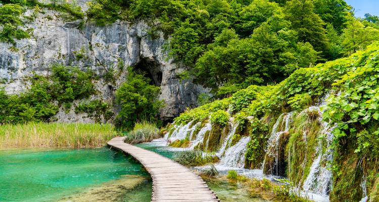 Wooden boardwalk over turquoise lake bordered by limestone cliffs and hanging greenery at Plitvice Lakes.