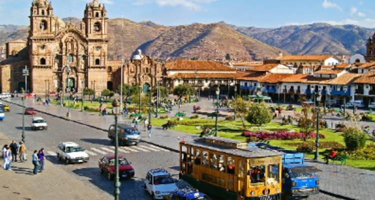Plaza de la ciudad concurrida con arquitectura histórica y montañas de fondo.