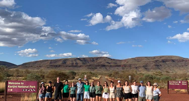 Large tour group standing in a line beneath the Welcome to Karijini National Park sign with hills behind.