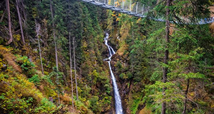 Hängebrücke über einen Wasserfall in einer bewaldeten Schlucht.