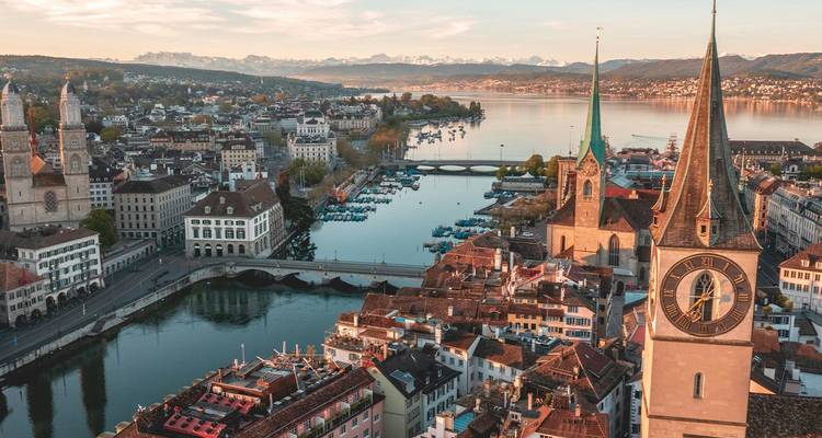 Aerial golden-hour panorama of Zurich’s Old Town, twin church spires, lake and distant snowy Alps.