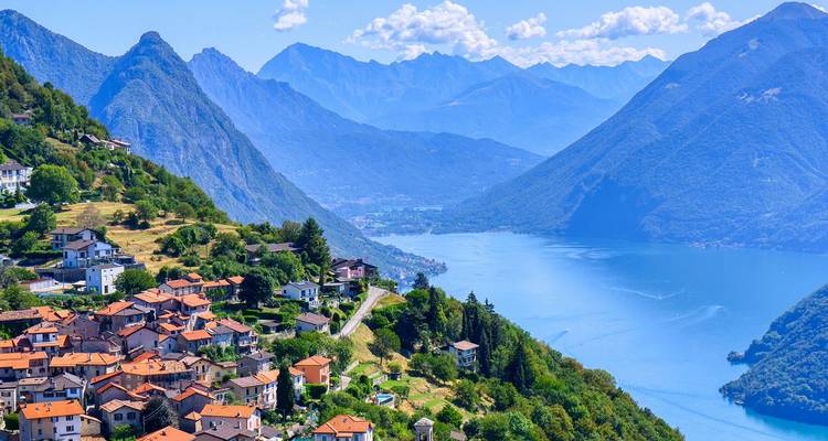 Bright blue Lake Lugano winding between steep green mountains with terracotta-roof villages on the slopes.