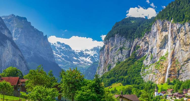 Un valle verde en Lauterbrunnen con acantilados alpinos escarpados, picos nevados y casas alpinas dispersas bajo un cielo azul vibrante.