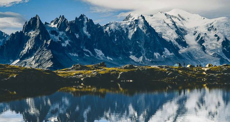 Jagged snow-covered alpine peaks reflected perfectly in a calm mountain lake under dramatic clouds.