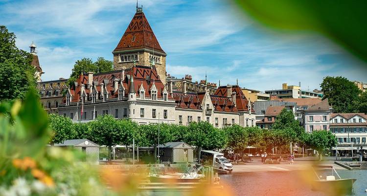 A picturesque lakeside chateau with red rooftops and turrets in Lausanne framed by leafy greenery and blue sky.