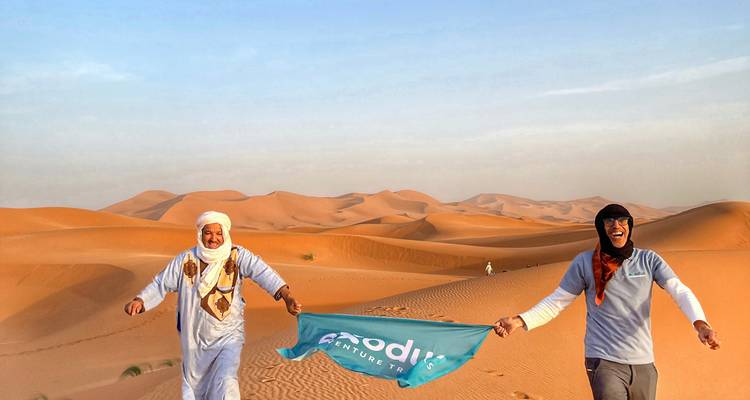 Two smiling guides holding a teal flag on rolling Sahara dunes with clear blue sky.