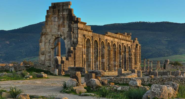 Ancient Roman stone arches of Volubilis ruins set against green hills in evening light.