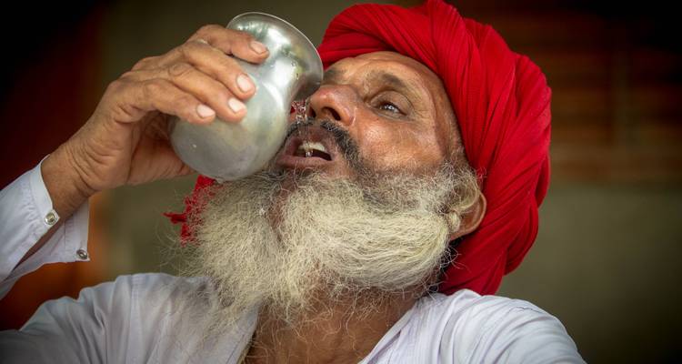 Man drinking water from a traditional metal jug with a red turban.