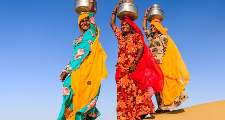 Three women in colorful outfits carrying pots on their heads across a desert landscape.