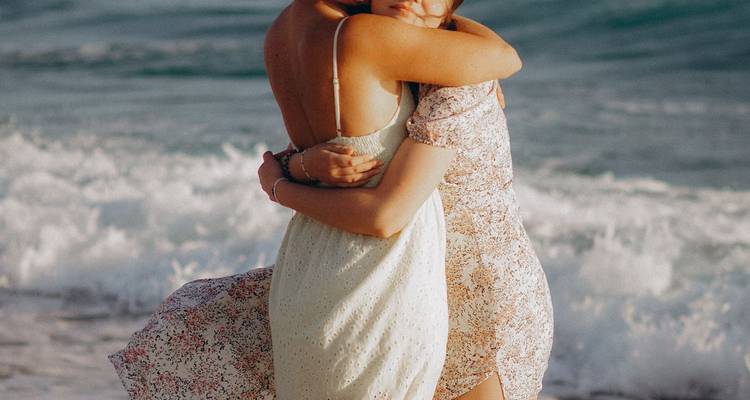 Two women embracing on a beach with waves in the background.