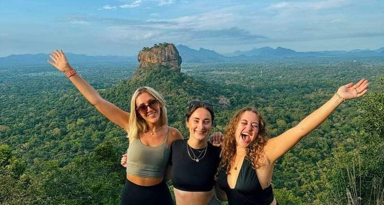 Three women celebrating with arms raised in front of a scenic viewpoint.