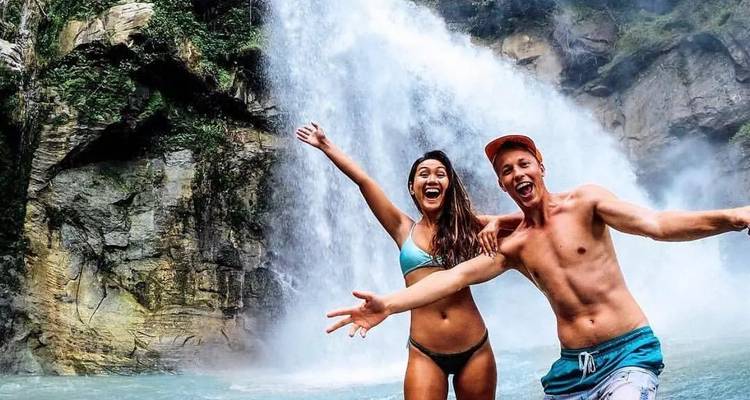 Two people enjoying a waterfall, arms raised, in swimwear.