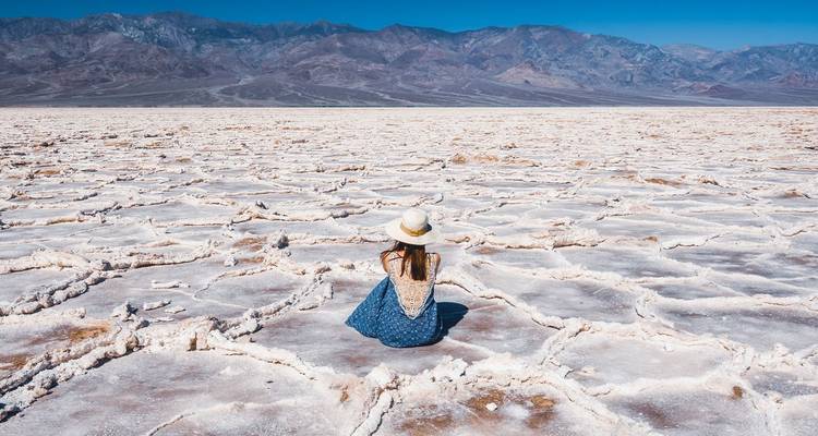 Jonge vrouw met een hoed zittend op de uitgestrekte gebarsten zoutplains van Death Valley met bergen daarachter
