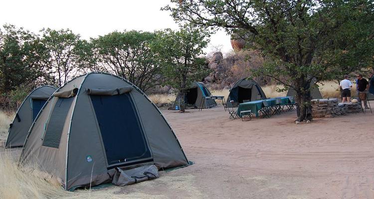Einfache Kuppelzelte und eine provisorische Essensecke befinden sich auf einem sandigen Buschcampingplatz unter Akazienbäumen in Botswana.