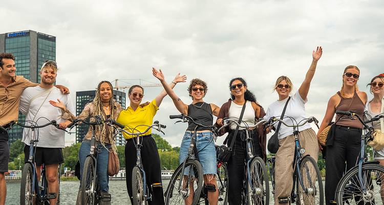 Group of friends on rental bicycles waving beside an urban canal with modern buildings behind.