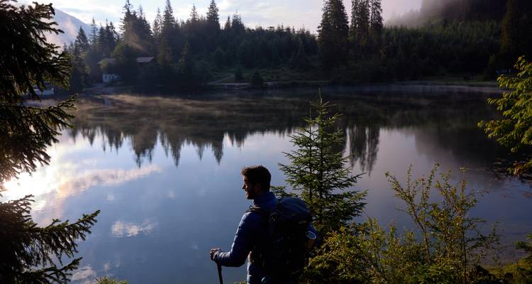 Person mit einem Rucksack, die über einen nebligen See am frühen Morgen blickt.