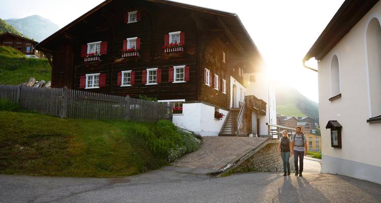 Zwei Menschen, die eine Straße mit traditionellen Häusern entlanggehen, während Sonnenlicht hindurchscheint.