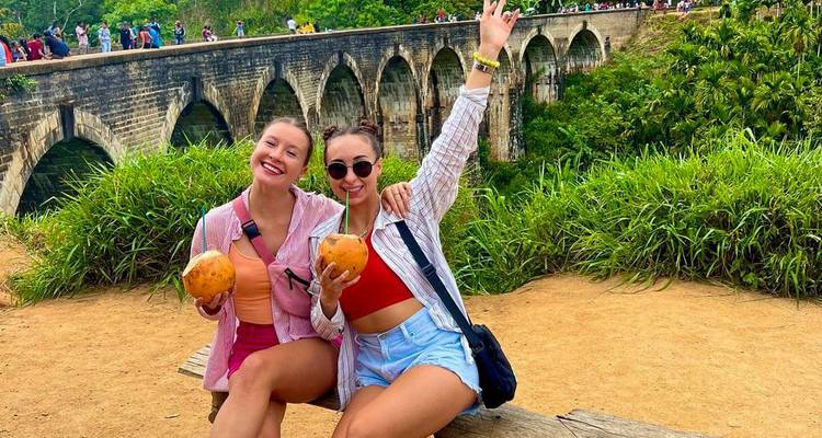 Deux femmes savourant des noix de coco avec un grand pont en arc de pierre en arrière-plan.