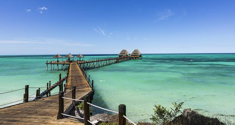 Wooden pier leading to thatched huts over clear blue water.