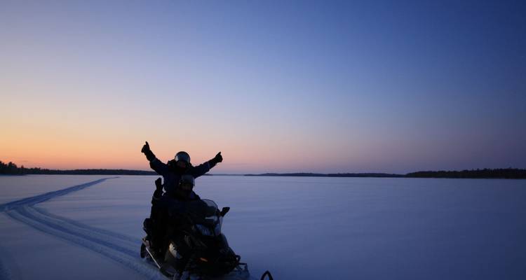 People riding a snowmobile across a snowy landscape at sunset.