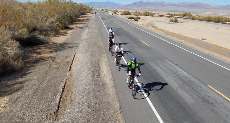 Group of cyclists riding on a desert road with a mountain range in the background.