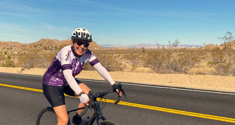 Cyclist smiling at the camera while riding on a road with desert scenery.
