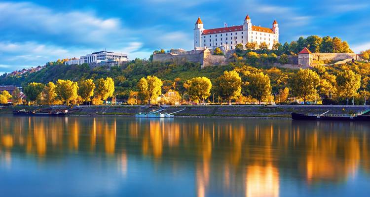 Le château de Bratislava au-dessus du Danube avec des arbres se reflétant dans l'eau.