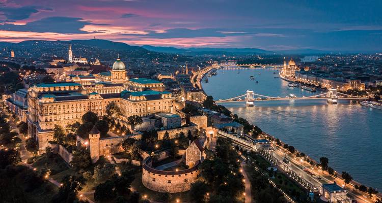 Aerial view of a city with a prominent river and illuminated buildings during sunset.