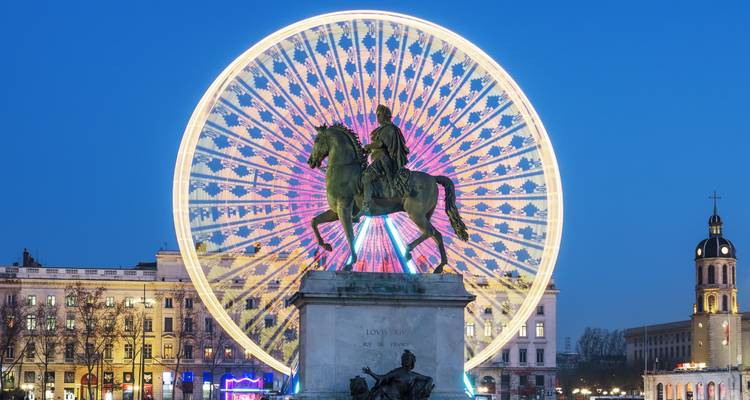 Noria iluminada y colorida girando detrás de una estatua ecuestre en una plaza urbana al anochecer.