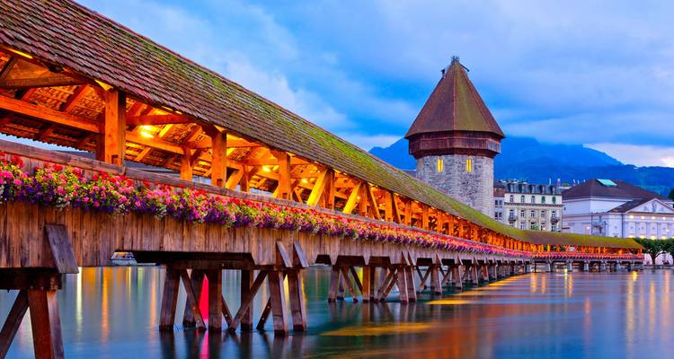 Puente de madera cubierto adornado con flores y torre de agua de piedra resplandeciendo sobre las aguas tranquilas del lago al crepúsculo.