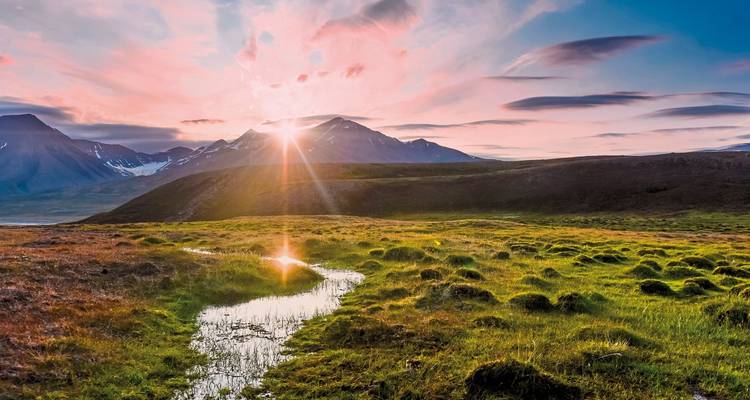 Coucher de soleil sur un paysage herbeux avec un ruisseau menant à un arrière-plan montagneux.