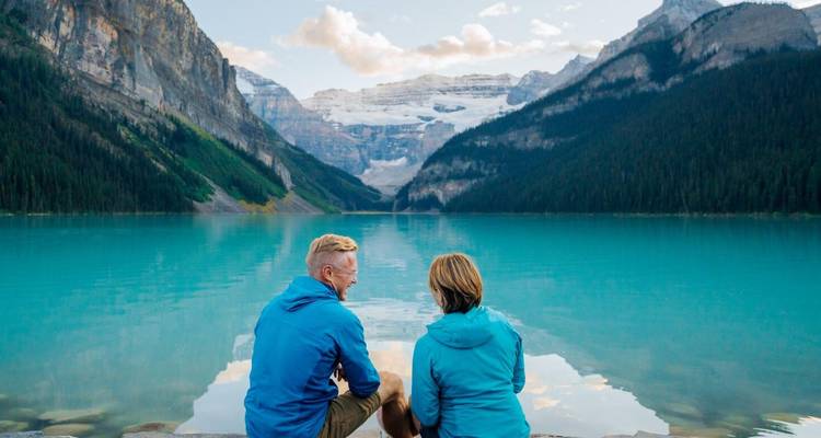 Deux personnes assises au bord des eaux claires du lac Louise avec des montagnes en arrière-plan.