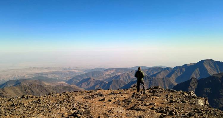 Wandelaar staat op een hoge rotsachtige bergtop van het Atlasgebergte en kijkt uit over uitgestrekte mistige valleien.
