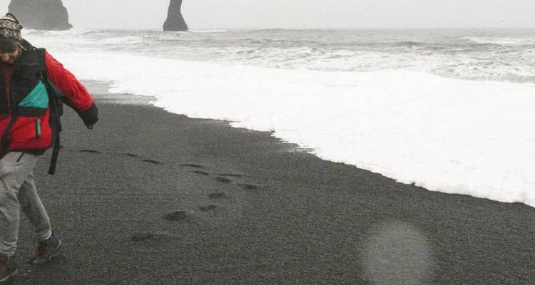 Hiker in red jacket walks along a black-sand beach with sea stacks looming through mist and surf.