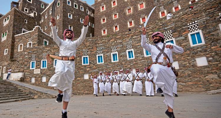Men performing traditional dance in front of stone buildings.