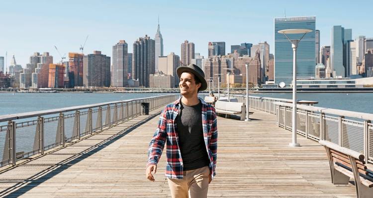 Lächelnde Person, die auf einem Pier mit der Skyline von New York City im Hintergrund geht.