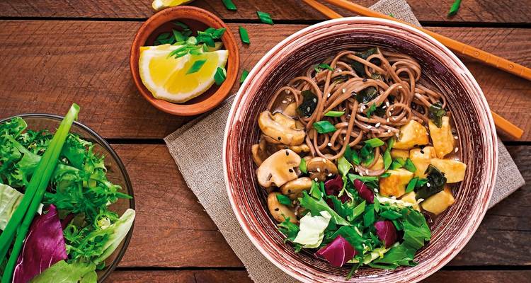 A bowl of noodle soup with mushrooms and vegetables on a wooden table.