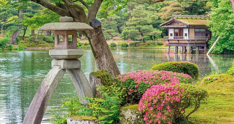 Ruhiger japanischer Garten mit steinerner Laterne und blühenden Sträuchern neben einem ruhigen Teich und einem hölzernen Teehaus.