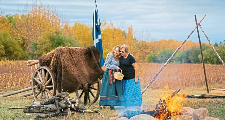 Two women dressed in traditional prairie attire posing by a campfire and ox-hide cart.