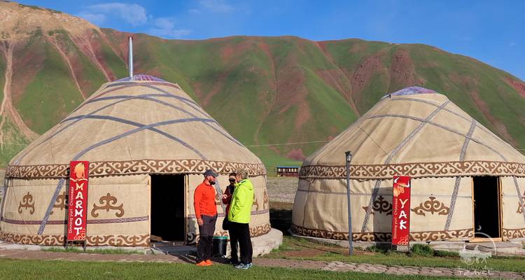 Deux personnes debout près de yourtes traditionnelles avec un paysage de montagnes en arrière-plan.